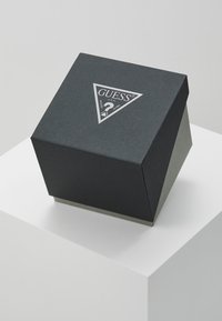 Boîte triangulaire noire avec surface texturée, présentant un logo argenté et un point d'interrogation sur le dessus. La base est d'un gris mat contrastant.