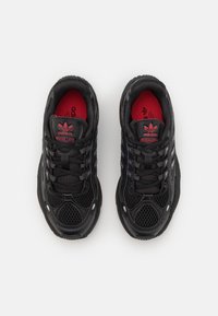 Black sneakers featuring a mesh upper, contrasting red insoles, and three signature stripes along the sides. Rounded, textured sole.
