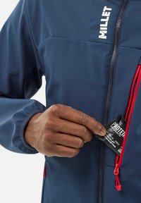 Veste softshell bleu marine avec une poche zippée sur la poitrine et des tirettes de zip rouges. Une main s'y introduit, révélant un emballage de collation protéinée noire.