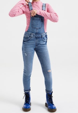 Personne ajustant les bretelles d’une salopette en denim bleu, portant un chemisier rose à manches longues et des bottes bleues à lacets sur fond blanc.