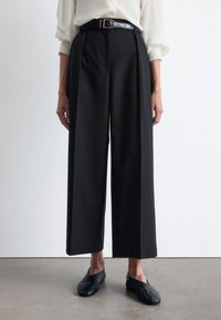 Black wide-leg trousers, featuring a high waist, front pleats, and a belt with a silver buckle. Smooth fabric and tailored design.