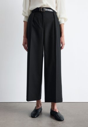 Schwarze weite Hosen mit hohem Bund, vorderen Falten und einem Gürtel mit silberner Schnalle. Glatte Stoffqualität und taillierte Passform.