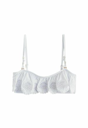 Kobieta z kręconymi czarnymi włosami w białym bikini, stojąca tyłem na tle słonecznej beżowej ściany.