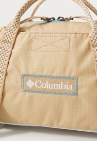 Beige textiele sporttas met blauwe accenten, voorzien van een gestikte logolabel en geweven handvatten, met een nadruk op een gladde textuur en een duurzaam ontwerp.