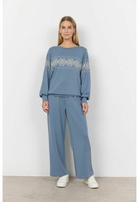 Blauwe loungewear set van zachte stof. Langsleeve top met decoratief wit sneeuwvlokkenpatroon; losse pasvorm, wijde broekspijpen. Witte sneakers.