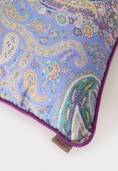 Coussin carré avec des motifs paisley et floraux pastel complexes sur un fond violet clair, fini avec un passepoil violet et une étiquette de marque en tissu.