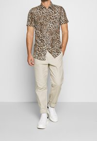 Mand iført kortærmet skjorte med leopardmønster, beige bukser med opsmøg og hvide sneakers med sort swoosh, går med den ene hånd i lommen.