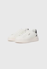 Hvite sneakers med en glatt lær overdel, rund tå, tykk gummisåle, og svart detalj ved hælen. Har snøring og logo detaljer.