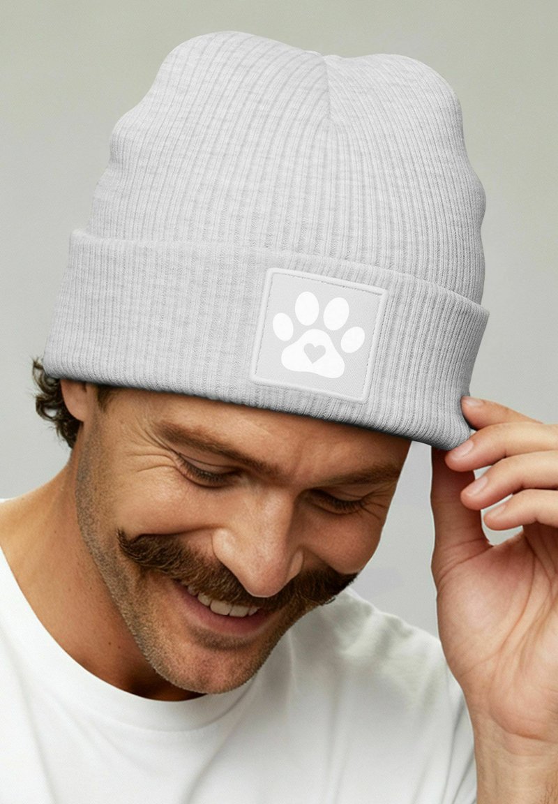 Man smiling and adjusting light gray knit beanie with paw print and heart logo on front, wearing white shirt against plain light background.