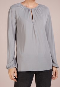 Hellgraue Bluse mit langen Ärmeln, einem Schlüsselloch-Ausschnitt, gerafften Details an den Schultern und gesicherten Manschetten. Glatte Stoffstruktur.
