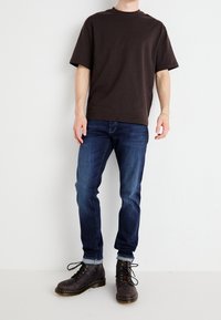 T-shirt a maniche corte marrone abbinato a jeans blu scuro e stivali marroni con lacci. L'outfit presenta una vestibilità rilassata e un design semplice.