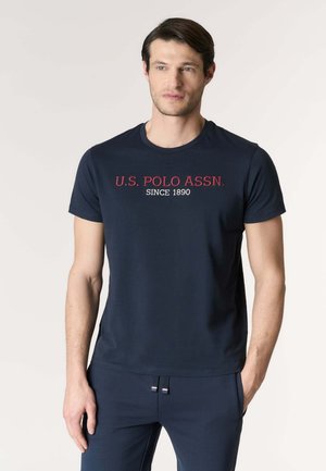 Uomo che indossa una maglietta blu navy con la scritta "U.S. POLO ASSN. SINCE 1890" in rosso e bianco, abbinata a pantaloni blu navy coordinati.