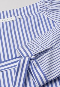 Chemise à rayures bleues et blanches avec un détail de manchette nouée, une texture lisse et un col classique. Étiquette ETERNA visible.