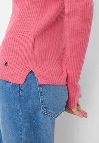 Aansluitende roze geribbelde trui met lange mouwen, zijnaad bij de zoom, gecombineerd met lichtblauwe denim. Klein logo nabij de onderkant links.