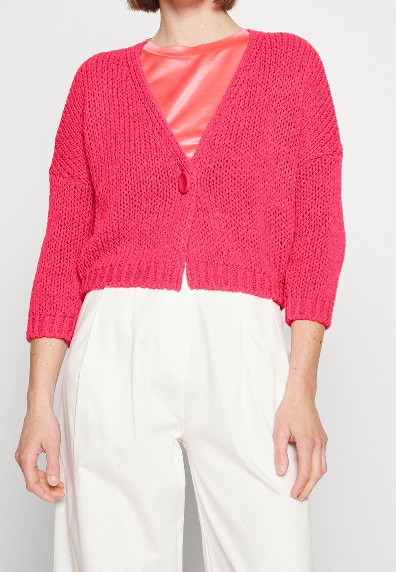 Růžový hrubě pletený cardigan s výstřihem do V, tříčtvrtečními rukávy a zapínáním na jeden knoflík. Nosit přes lesklý korálový top a bílé kalhoty.