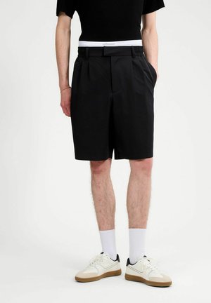 Persoon draagt zwarte geplooide korte broek, witte enkelsokken en witte sneakers met bruine zolen, staand tegen een effen witte achtergrond.