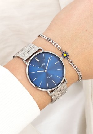 Montre en acier inoxydable argenté avec un cadran rond bleu et des aiguilles argentées. Elle possède un bracelet en maille et un bracelet chaîne avec un accent floral.