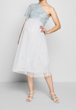 Vrouw draagt een witte jurk met één schouder, een zilveren top met pailletten en een geplooide rok, poseert met één hand op haar heup en witte sandalen met bandjes.