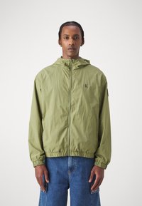 Giacca leggera di colore verde chiaro, con cappuccio, chiusura a zip frontale e polsini elasticizzati. Presenta un logo sul petto e una vestibilità comoda.