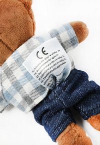 Ursuleț de pluș îmbrăcat într-o cămașă în carouri albăstrui și gri, pantaloni albaștri din denim, cu o etichetă de îngrijire vizibilă pe spate. Blană maro pe lăbuțe și față.