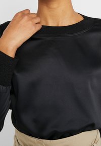 Person klädd i en svart satin långärmad topp med ribbade manschetter och krage, instoppad i beige högmidjade byxor, hand nära kragen.