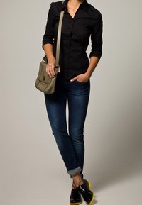 Camicia nera con bottoni, jeans blu scuro arrotolati sui polsini e scarpe nere lucide con suole gialle. Borsa a tracolla grigia indossata a corsa.