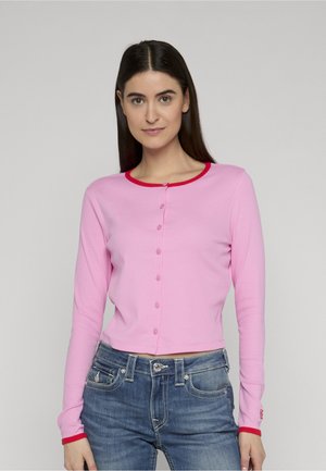 Pikad tumedad juuksed omav naine kannab roosat nööpidega kaunistatud kardigani, millel on punane ääris, ja siniseid teksaseid, seistes ühetaolise helehalli tausta ees.