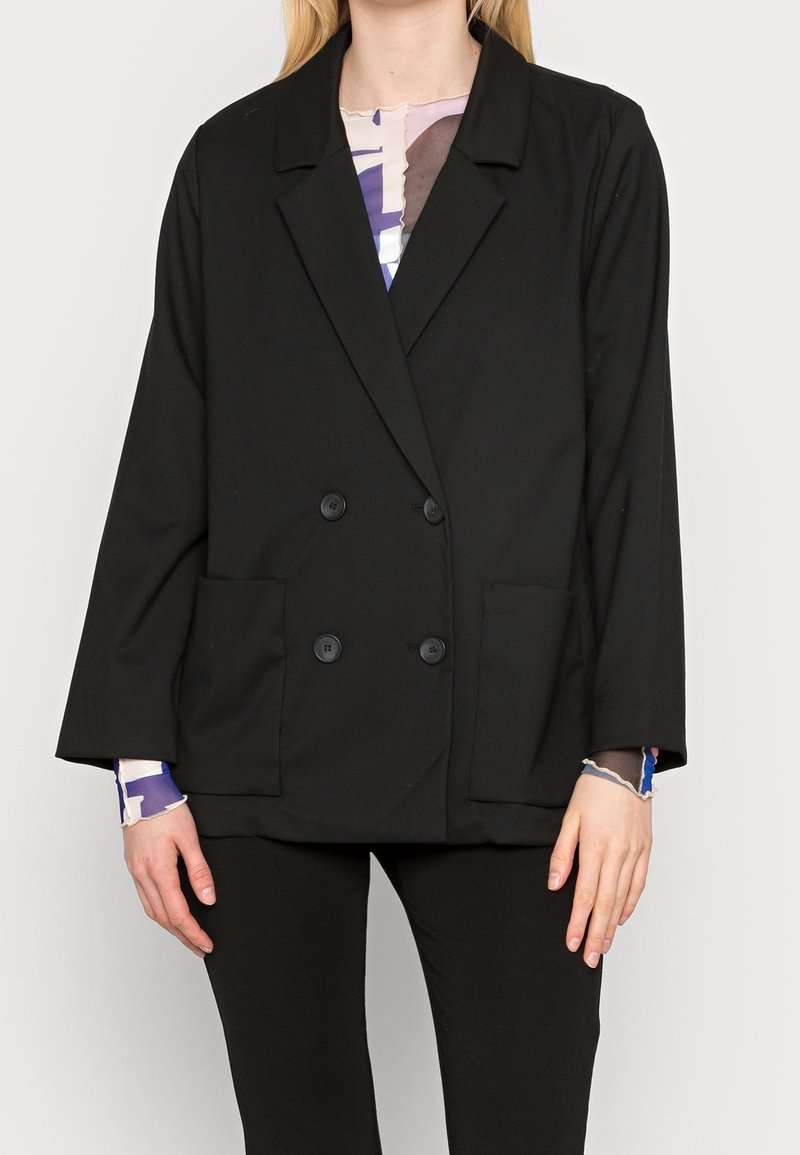 Femme portant un blazer noir croisé avec deux poches avant sur un haut à manches longues à motifs, associé à un pantalon noir.