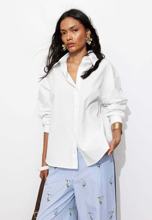 Femme aux longs cheveux foncés portant une chemise blanche ample boutonnée, un pantalon bleu à jambes larges avec motif floral, des boucles d'oreilles en or et un bracelet en or.