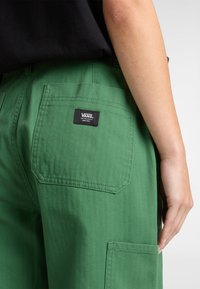 Groene cargobroek van textuurstof, met een achterzak met een zwart Vans-logo label, gepresenteerd op een model van opzij.