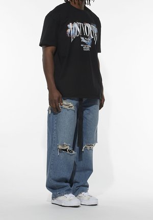 Czarny grafitowy t-shirt z białym napisem, w połączeniu z jasnoniebieskimi przetartymi dżinsami z dziurami; noszony z białymi sneakersami.