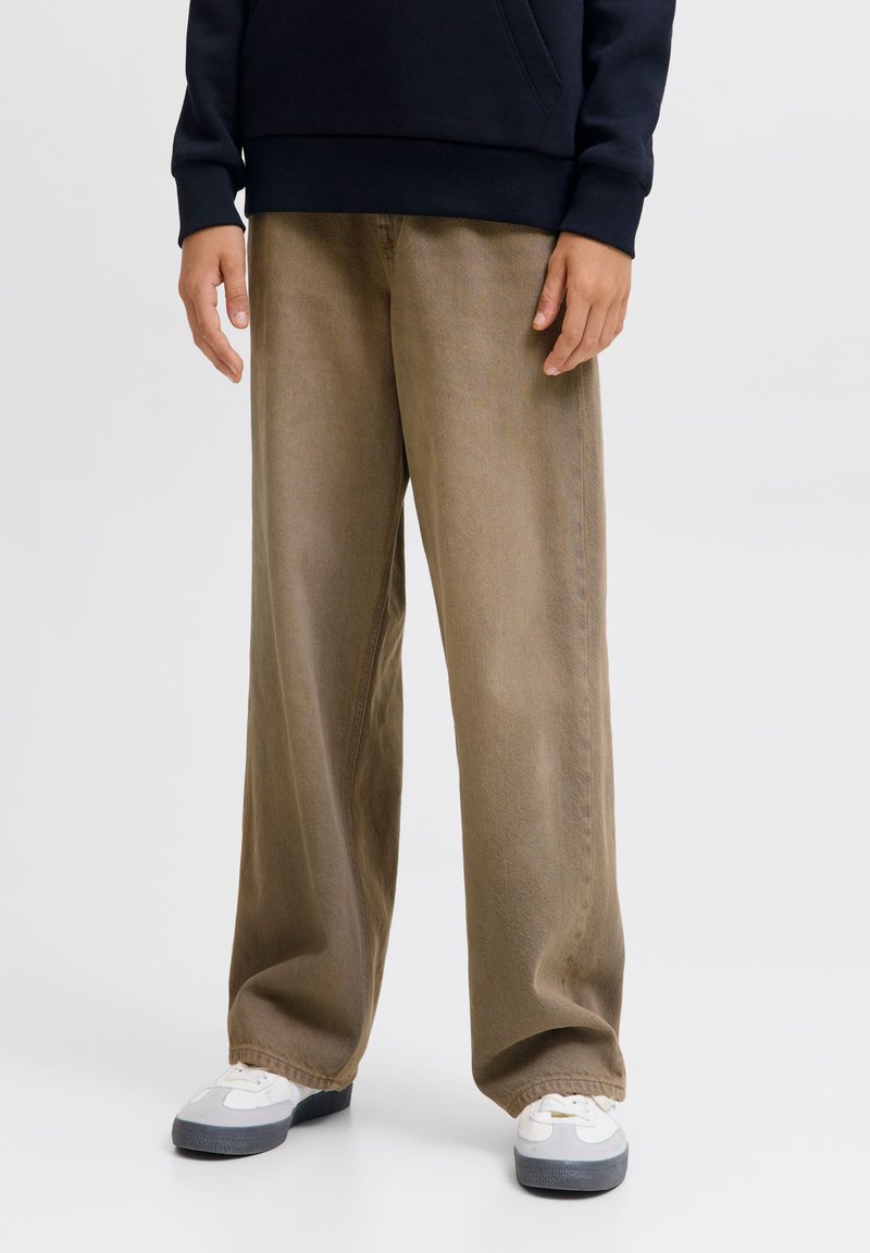 Personne portant un pantalon marron ample, des baskets blanches et grises, et un haut à manches longues foncé, debout devant un fond uni.