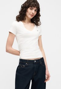 Γυναίκα με σγουρά μαλλιά φοράει εφαρμοστό λευκό T-shirt με V-λαιμόκοψη και σκουρόχρωμο μπλε τζιν, στέκεται με το ένα χέρι πίσω από την πλάτη της.