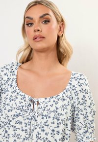 Blusa bianca con stampa floreale blu, scollatura quadrata e maniche a sbuffo. Presenta un dettaglio a fiocco sul davanti, texture arricciata e una leggera lucentezza.