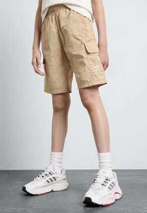Personne portant un short cargo beige avec des poches latérales et des baskets blanches avec des détails noirs et rouges sur un sol gris.