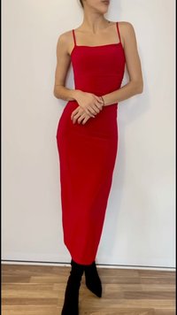 Vrouw die een nauwsluitende rode midi-jurk met dunne bandjes draagt, staand met gekruiste handen en zwarte enkellaarsjes op een houten vloer.