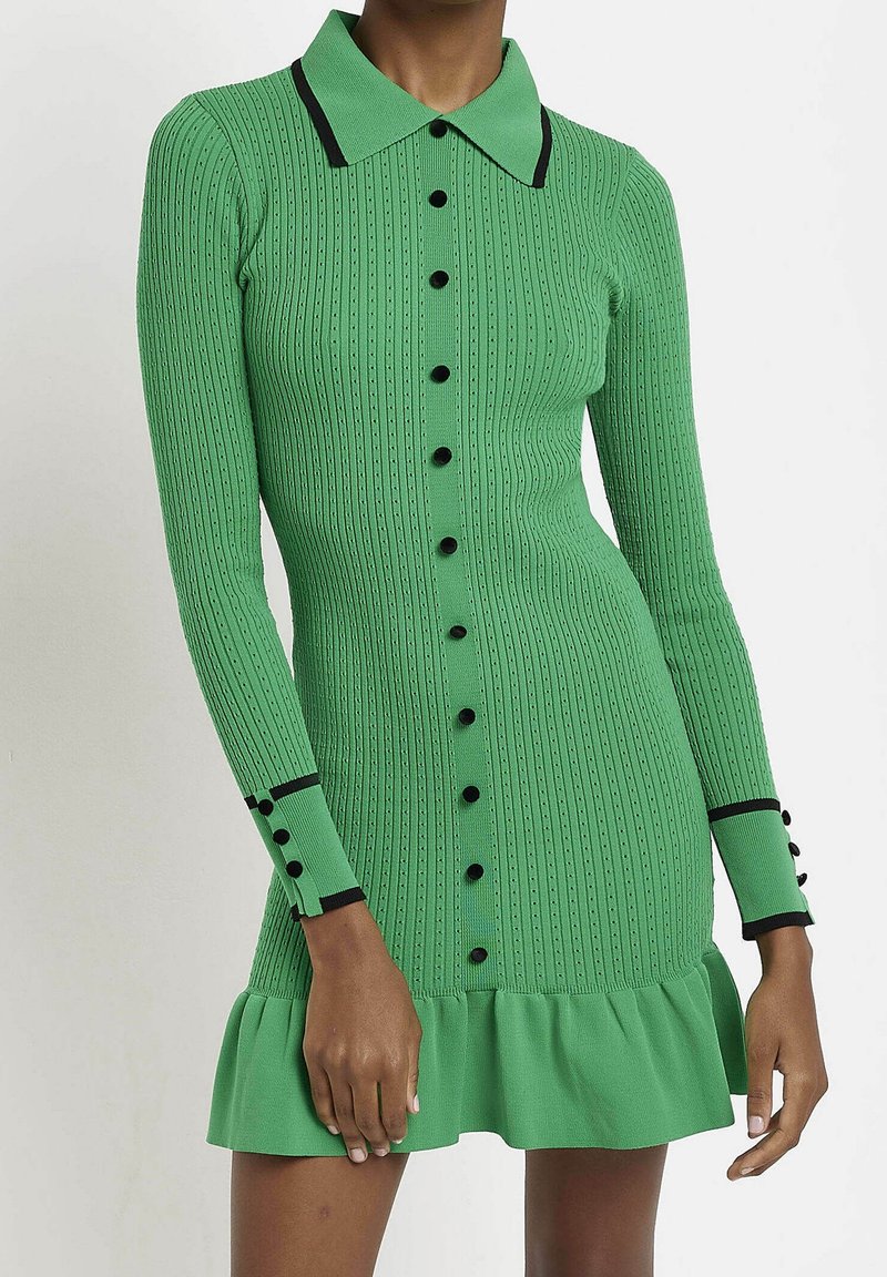 Robe verte à côtes avec un col, accents de boutons noirs, manches longues et ourlet volanté. Tissu texturé avec des lignes verticales.