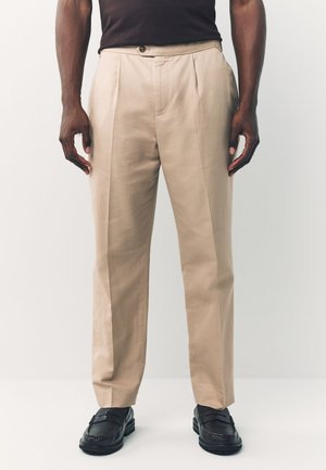Pantaloni beige plissettati con chiusura a bottoni e tasche laterali, realizzati in tessuto liscio, abbinati a mocassini neri.