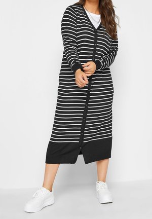 Femme portant une robe cardigan à boutons rayée horizontale noire et blanche avec des baskets blanches, ajustant le poignet de la manche, sur fond uni.