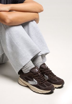 Person sitting cross-legged wearing light gray pants, ribbed gray socks, and dark brown athletic sneakers with beige soles and silver accents.