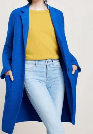 Vrouw die een felblauwe jas draagt over een mosterdgele trui en lichtblauwe spijkerbroek, handen in de jaszakken, staand tegen een effen achtergrond.