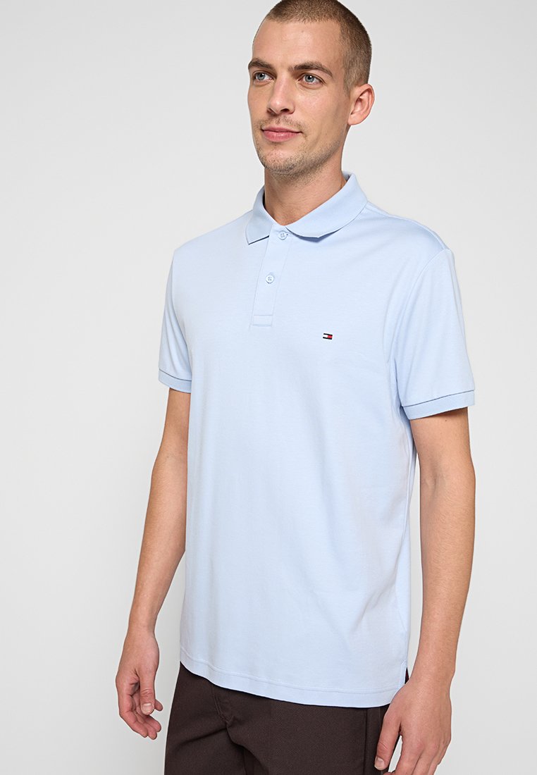 Muž v světlomodrém polokošili s malým logem na hrudi, stojící na neutrálním světlém pozadí.