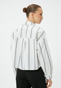 White long-sleeve shirt with black vertical stripes, featuring a back pleat detail, classic collar, and buttoned cuffs.