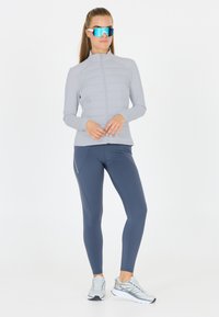 Lysegrå jakke med lynlås og kviltede sektioner, parret med mørkegrå leggings. Tilbehøret inkluderer overdimensionerede reflekterende solbriller og grå træningssko.