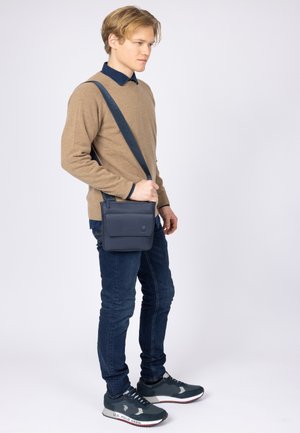 Borsa a tracolla in pelle blu navy con patta frontale e tasca con zip, indossata sopra un maglione beige e jeans blu, con scarpe da ginnastica grigie.