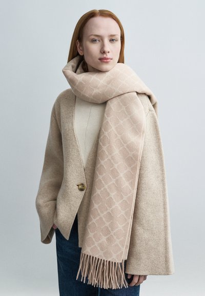 Γυναίκα με κόκκινα μαλλιά που φοράει μπεζ ζακέτα, μπεζ κασκόλ με σχέδια και μπλε τζιν παντελόνι, στέκεται μπροστά σε ένα απλό ανοιχτό γκρι φόντο.