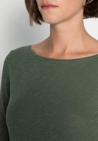 Groene lange mouw top met een ronde hals. De stof heeft een subtiele gemêleerde textuur en een getailleerd silhouet.