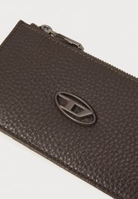 Brown leather wallet with a textured surface, featuring a dark metal logo accent and zip closure. Smooth edges and stitching details visible.