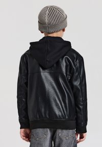 Bambino che indossa una giacca in finta pelle nera con cappuccio, jeans in denim grigi e un berretto lavorato a maglia beige e nero a righe, in piedi di spalle.