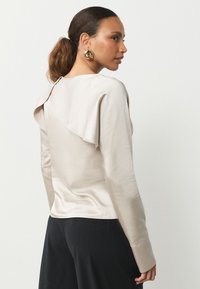 Camisa beige claro de manga larga con un diseño drapeado en la espalda y textura suave. Combinada con pantalones negros que cuentan con puños anchos.